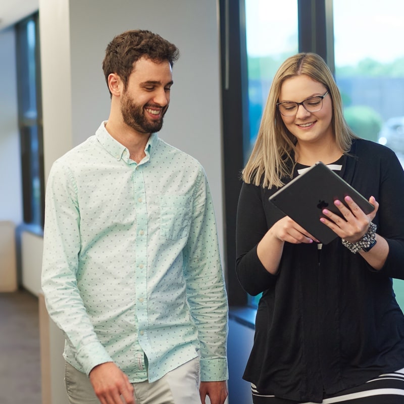 A young man and a woman walking through a hallway while looking at an iPad the woman is holding