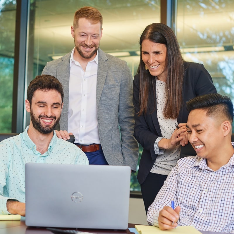 Four young professionals in business casual attire gathering around a laptop