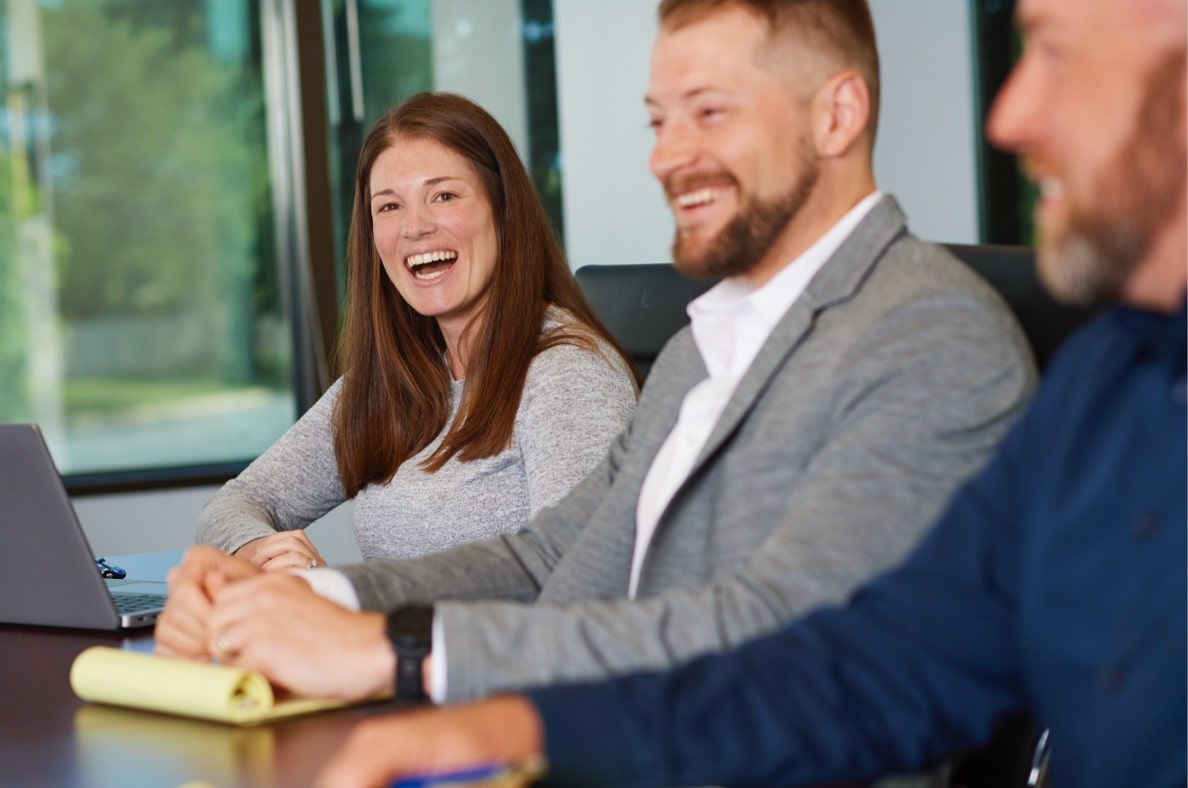 A woman and two men in professional attire at a meeting table with a laptop and legal pad